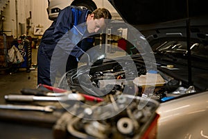 Young concentrated garage mechanic, technician, car engineer holding a flashlight lamp and repairing car under hood in the repair