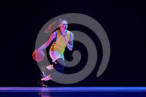 Young concentrated girl, basketball player in motion, running with ball against black studio background in neon light