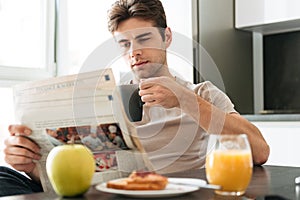 Young concentrated man reading newspaper while sitting in kitchen