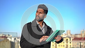 Young concentrated man surfing social media on tablet looking away thinking standing outdoors. Portrait of focused