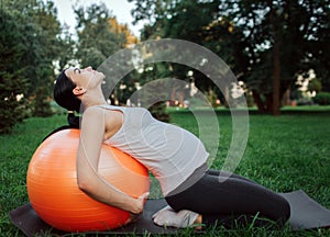 Young concentrated pregnant woman sit on knees on yoga mate in park. She lean to orange fitness ball with back and look