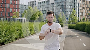 A young concentrated sportive man does morning jog on the sidewalk and checks the time on the watch. An athlete guy