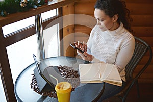 Young concentrated woman checking her mobile phone while sitting near window at wooden cafe