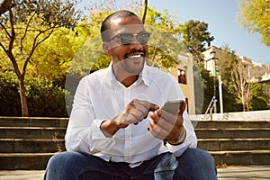 Young confident african man pointing hand on smartphone while sitting at sunny city park.Concept of happy business