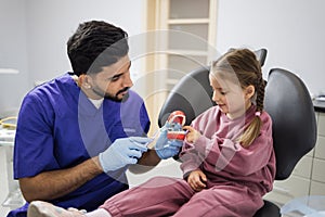 Young confident bearded pediatric dentist showing to little preschool girl how to brush teeth