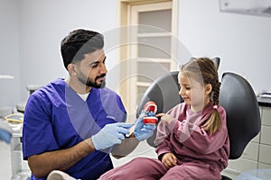 Young confident bearded pediatric dentist showing to little preschool girl how to brush teeth