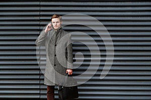 Young confident businessman in a gray coat and briefcase in his hand is talking on mobile phone against wall of modern