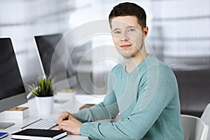 Young confident businessman, wearing a green pullover, is sitting at the desk in a modern office, while working on the