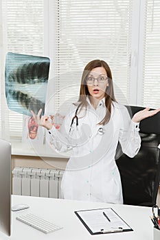 Young confident female doctor standing at desk, holding X-ray of lungs, fluorography, roentgen in light office in