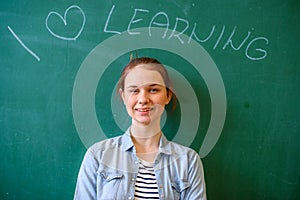 Young confident female high school student standing in front of chalkboard in classroom, looking at camera and smiling.