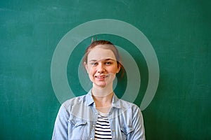 Young confident female high school student standing in front of chalkboard in classroom, looking at camera and smiling.