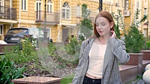 Young confident ginger woman in jacket walking down street in the city, yellow building background