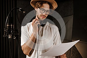 Young confident male singer thoughtfully reading text of new song working in sound recording studio