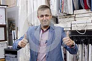 Young confident positive businessman fabric store owner showing thumbs up, gesture sign ok. Man standing in the store background