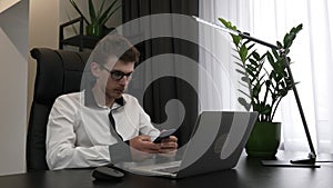 Young confident smart businessman is using smartphone sitting in his modern office desk. Caucasian male worker with mobile phone i