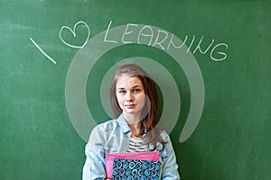 Young confident smiling female high school student standing in front of chalkboard in classroom, holding textbooks.