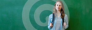 Young confident smiling female high school student standing in front of chalkboard in classroom, wearing a backpack.