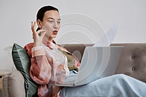 Young confident tutor with paper sitting on couch with laptop on her knees