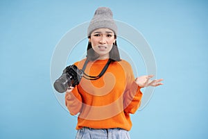 Young confused asian girl in hat, holds digital camera and shrugging shoulders, clueless how to take pictures on digicam