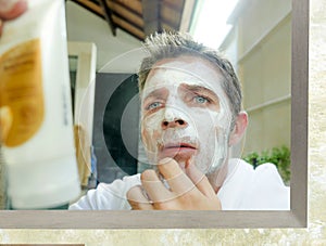 Young confused Caucasian man at home reading instructions of white facemask product while applying the facial mask on his face