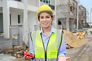 Young construction engineer woman in safety vest with yellow helmet holding radio, standing on building construction site. Smiling