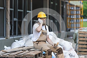 young construction worker in hardhat and protective googles talking on smartphone and having lunch