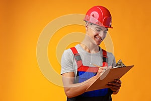 Young construction worker wearing hardhat and holding a clipboard on yellow background in studio