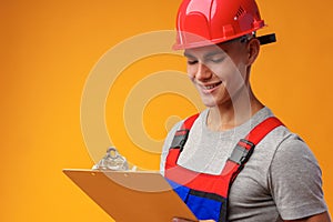 Young construction worker wearing hardhat and holding a clipboard on yellow background in studio