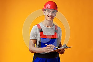 Young construction worker wearing hardhat and holding a clipboard on yellow background in studio