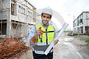 Young constructor engineer man smiling holding blueprint while talking about building construction, looking camera on under-