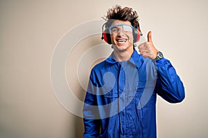 Young constructor man wearing uniform and earmuffs over isolated white background doing happy thumbs up gesture with hand