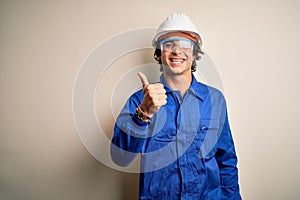 Young constructor man wearing uniform and security helmet over isolated white background doing happy thumbs up gesture with hand