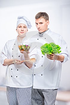 young cooks in suits with salad in hands on gray