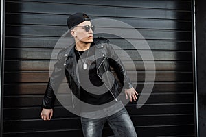 Young cool hipster man in stylish T-shirt in a trendy black baseball cap with dark sunglasses in a leather trendy black jacket