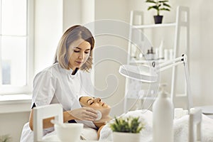 Young cosmetologist or dermatologist cleaning lying womans face with cotton pads in spa salon