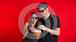 Young couple in 3d glasses watching fascinating comedy or horror movie and eating popcorn on red studio background.
