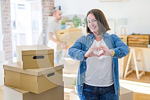 Young couple arround cardboard boxes moving to a new house, plus size woman standing at home smiling in love showing heart symbol