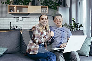 Young couple Asian man and woman at home sitting on sofa together using laptop, happy and smiling multiracial family looking at