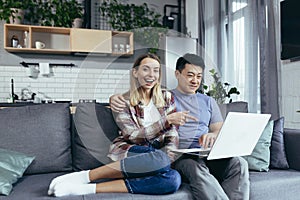 Young couple Asian man and woman at home sitting on sofa together using laptop, happy and smiling multiracial family looking at