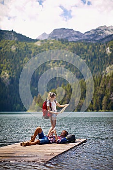 Young couple with bacpacks on adventure. Man lying on wooden jetty by lake, woman looking at map. Lifestyle, love, togetherness,