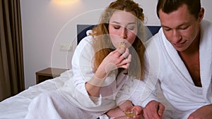 Young couple in bathrobes having a breakfast together in bed in hotel room.