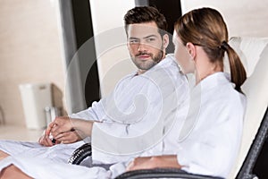 young couple in bathrobes holding hands and looking at each other while resting together