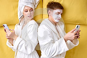 Young couple in bathrobes lying on bed using smartphones, suspecting each other