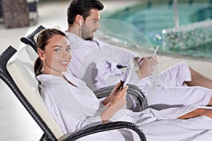 young couple in bathrobes using digital devices while resting near pool