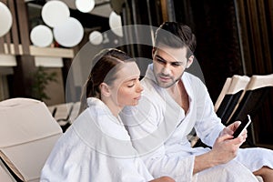 young couple in bathrobes using smartphone together in spa