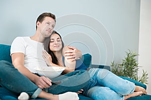 Young couple beautiful brunette woman and handsome man watching movie on the sofa eating pop corn hugging cozy lifestyle