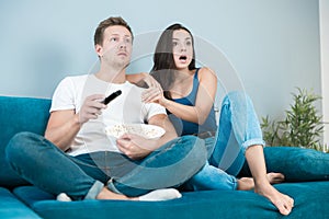 Young couple beautiful brunette woman and handsome man watching movie on the sofa eating pop corn looking surprised cozy