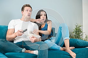 Young couple beautiful brunette woman and handsome man watching scary movie on the sofa eating pop corn looking