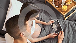 A young couple in bed and are looking at a picture of a baby on an ultrasound scan
