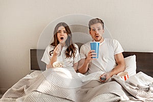 Young couple in the bed. A scared beautiful man and woman are eating popcorn and watching TV together in bedroom